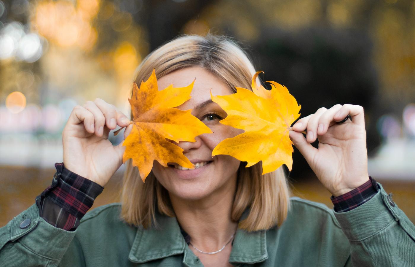 A smiling woman playfully holding two autumn leaves over her eyes