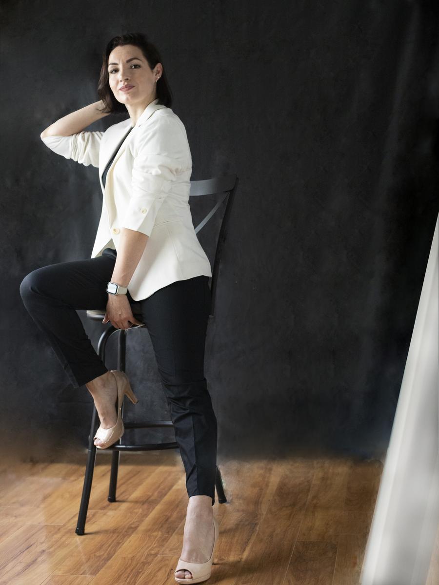 Professional woman in a white blazer posed on a stool against a dark backdrop