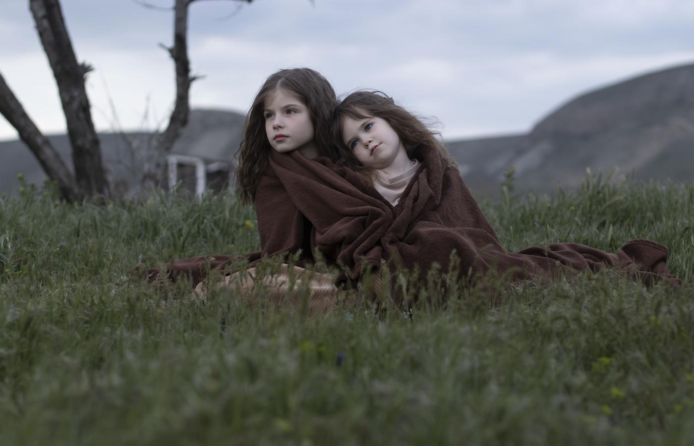 Two girls wrapped together in a brown blanket beneath a mountain sky